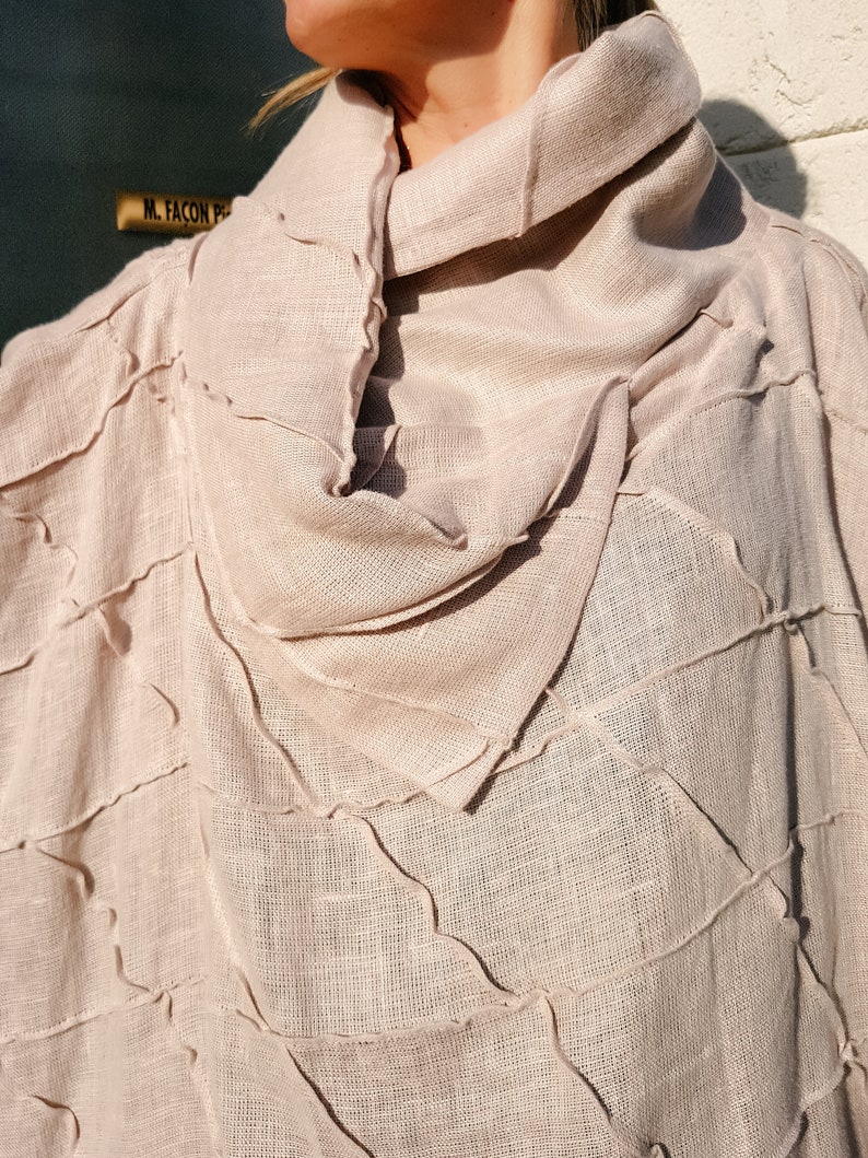 May include: A light beige linen poncho with a high, gathered neckline. The fabric has a textured, woven look and is stitched with a decorative, uneven seam.