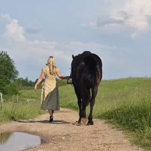 May include: A woman in a light-colored dress walks alongside a black horse on a dirt path. The horse is on the right, and the woman is on the left. The path has a puddle of water. Green grass and trees are in the background under a cloudy sky.