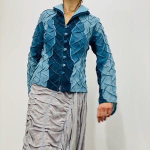 May include: A blue denim jacket with a textured, quilted design. The jacket has a collar and a button-up front. The model is wearing a light grey skirt.