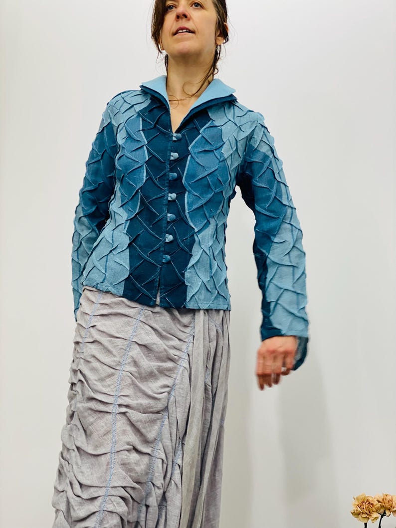 May include: A blue denim jacket with a textured, quilted design. The jacket has a collar and a button-up front. The model is wearing a light grey skirt.