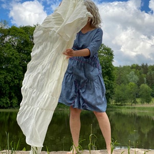 Puede incluir: Una mujer lleva un vestido de mezclilla azul con costuras rosas y una tela de lino blanca drapeada sobre su cabeza. Est&aacute; de pie sobre una roca junto a un lago con hierba verde y &aacute;rboles en el fondo.