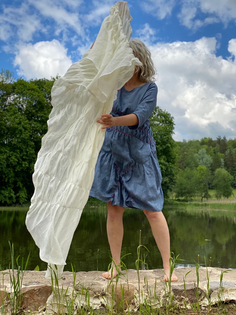 Puede incluir: Una mujer lleva un vestido de mezclilla azul con costuras rosas y una tela de lino blanca drapeada sobre su cabeza. Est&aacute; de pie sobre una roca junto a un lago con hierba verde y &aacute;rboles en el fondo.