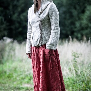 May include: A woman wearing a white hooded jacket with a floral pattern and a long, red, textured skirt. She is standing in a field of tall grass.