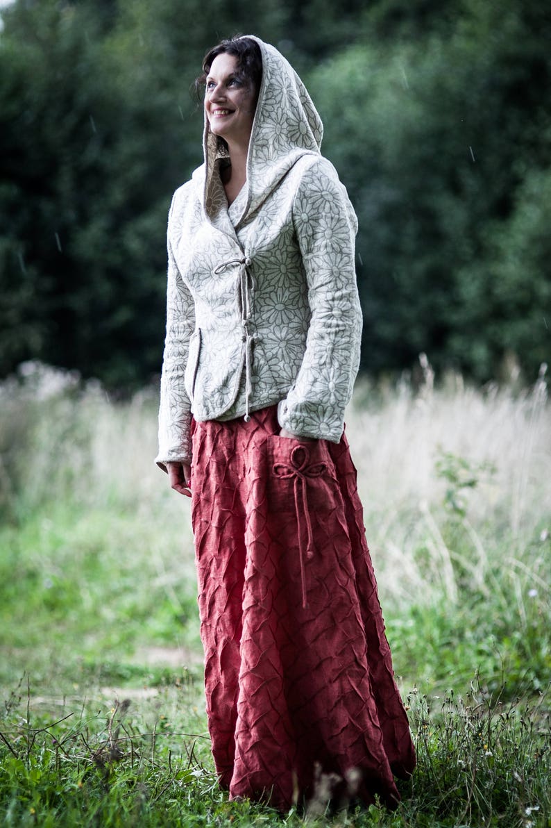 May include: A woman wearing a white hooded jacket with a floral pattern and a long, red, textured skirt. She is standing in a field of tall grass.