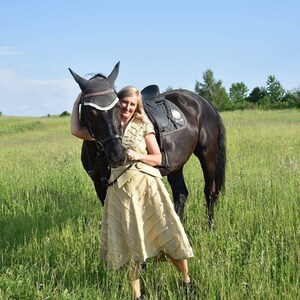 May include: A woman stands next to a black horse in a grassy field under a blue sky. The horse has a saddle and bridle. The woman wears a tan skirt and top. The scene evokes a sense of equestrian lifestyle and outdoor activity.