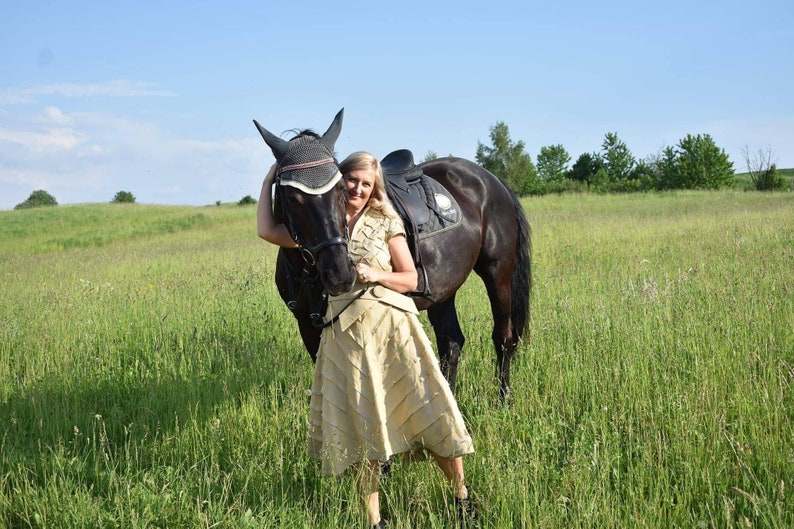 May include: A woman stands next to a black horse in a grassy field under a blue sky. The horse has a saddle and bridle. The woman wears a tan skirt and top. The scene evokes a sense of equestrian lifestyle and outdoor activity.