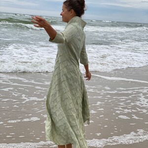 May include: A woman wearing a long, light green dress with a textured pattern stands on a sandy beach with waves crashing in the background.