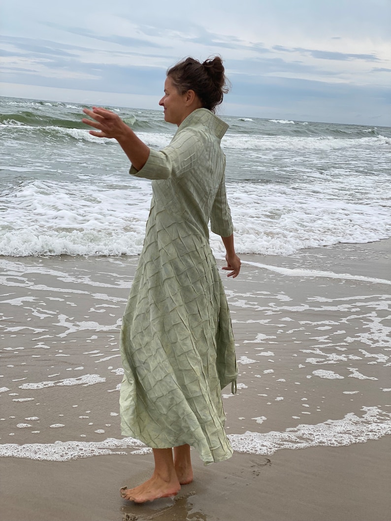 May include: A woman wearing a long, light green dress with a textured pattern stands on a sandy beach with waves crashing in the background.