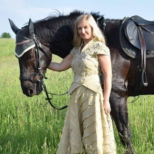 May include: A woman in a beige dress stands next to a black horse in a grassy field. The dress has a layered design and a matching belt. The horse wears a bridle and saddle, suggesting equestrian activities.