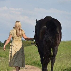May include: A woman in a beige dress walks alongside a black horse on a dirt path. The horse is wearing a bridle. The scene takes place on a sunny day with a blue sky and green fields in the background.