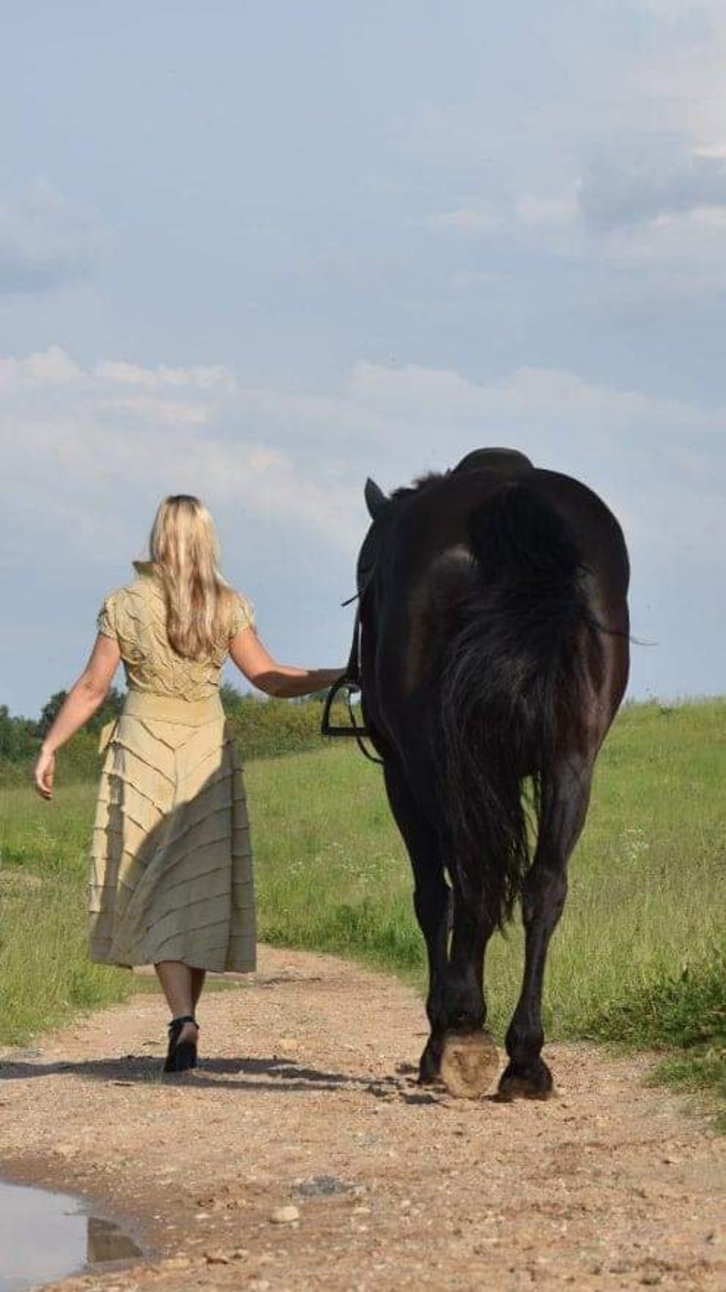 May include: A woman in a beige dress walks alongside a black horse on a dirt path. The horse is wearing a bridle. The scene takes place on a sunny day with a blue sky and green fields in the background.