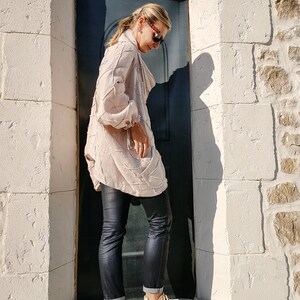 May include: A woman wearing a light pink, textured, long-sleeved shirt, black jeans, and beige platform sneakers. She is standing in front of a dark blue door.