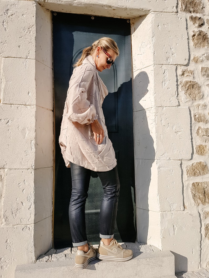 May include: A woman wearing a light pink, textured, long-sleeved shirt, black jeans, and beige platform sneakers. She is standing in front of a dark blue door.