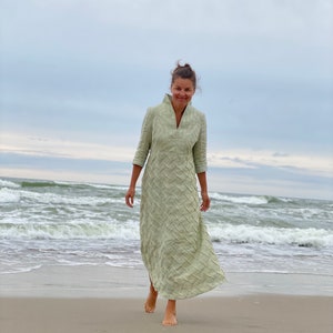May include: A woman wearing a light green, long-sleeved dress with a textured pattern walks on a sandy beach with waves in the background.