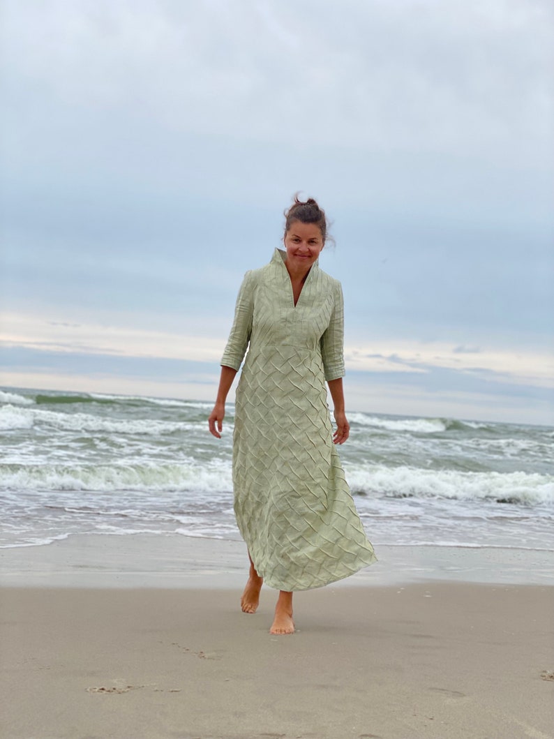 May include: A woman wearing a light green, long-sleeved dress with a textured pattern walks on a sandy beach with waves in the background.