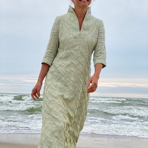 May include: A woman wearing a light green dress with a textured pattern stands on a sandy beach with waves in the background.