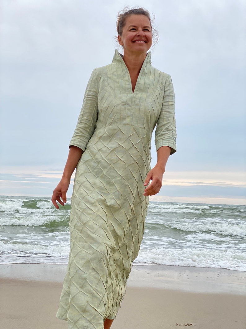 May include: A woman wearing a light green dress with a textured pattern stands on a sandy beach with waves in the background.