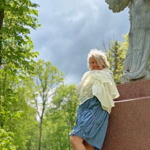 Puede incluir: Una mujer con un vestido azul claro y una chalina blanca de pie junto a una estatua de &aacute;ngel de piedra en un parque.