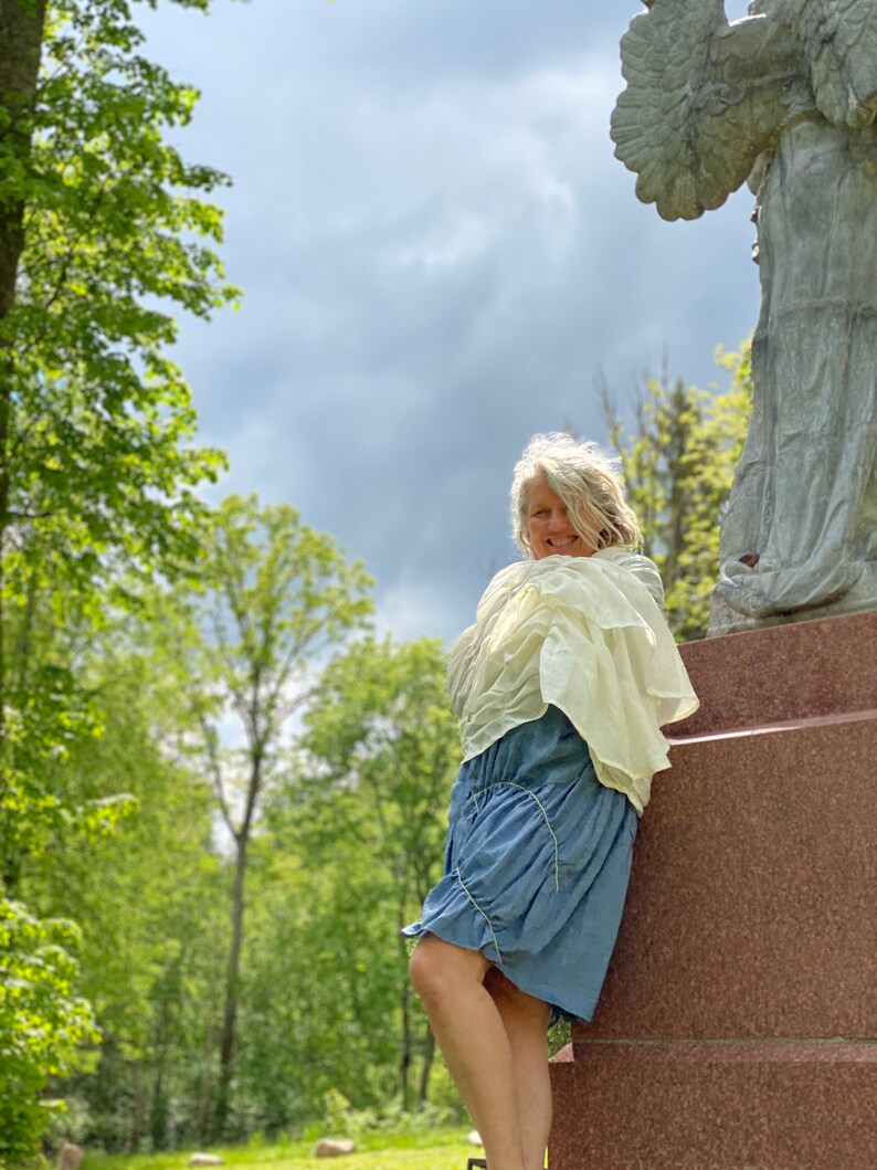 Puede incluir: Una mujer con un vestido azul claro y una chalina blanca de pie junto a una estatua de &aacute;ngel de piedra en un parque.