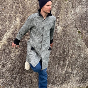 Puede incluir: Una chaqueta gris de manga larga con cuello y puños negros. La chaqueta tiene un patrón geométrico y está abotonada. La persona que lleva la chaqueta también lleva pantalones vaqueros azules y una boina negra.