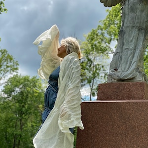 Puede incluir: Una mujer con una bufanda blanca transparente se encuentra de pie frente a una estatua de &aacute;ngel de piedra. La mujer lleva un vestido azul y tiene el pelo recogido hacia atr&aacute;s. La estatua est&aacute; sobre un pedestal de piedra.