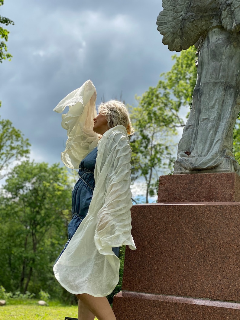 Puede incluir: Una mujer con una bufanda blanca transparente se encuentra de pie frente a una estatua de &aacute;ngel de piedra. La mujer lleva un vestido azul y tiene el pelo recogido hacia atr&aacute;s. La estatua est&aacute; sobre un pedestal de piedra.