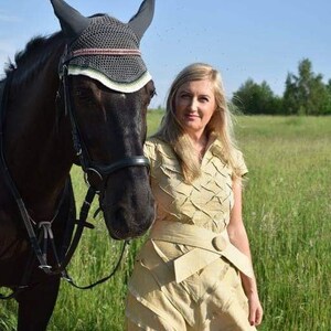 May include: A dark brown horse wearing a black and gray fly bonnet and bridle stands next to a person wearing a light yellow dress with a belt. The background is a green field and blue sky.