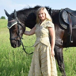 May include: A woman in a beige dress stands next to a black horse in a grassy field. The dress has a layered design and a matching belt. The horse wears a bridle and saddle, suggesting equestrian activities.