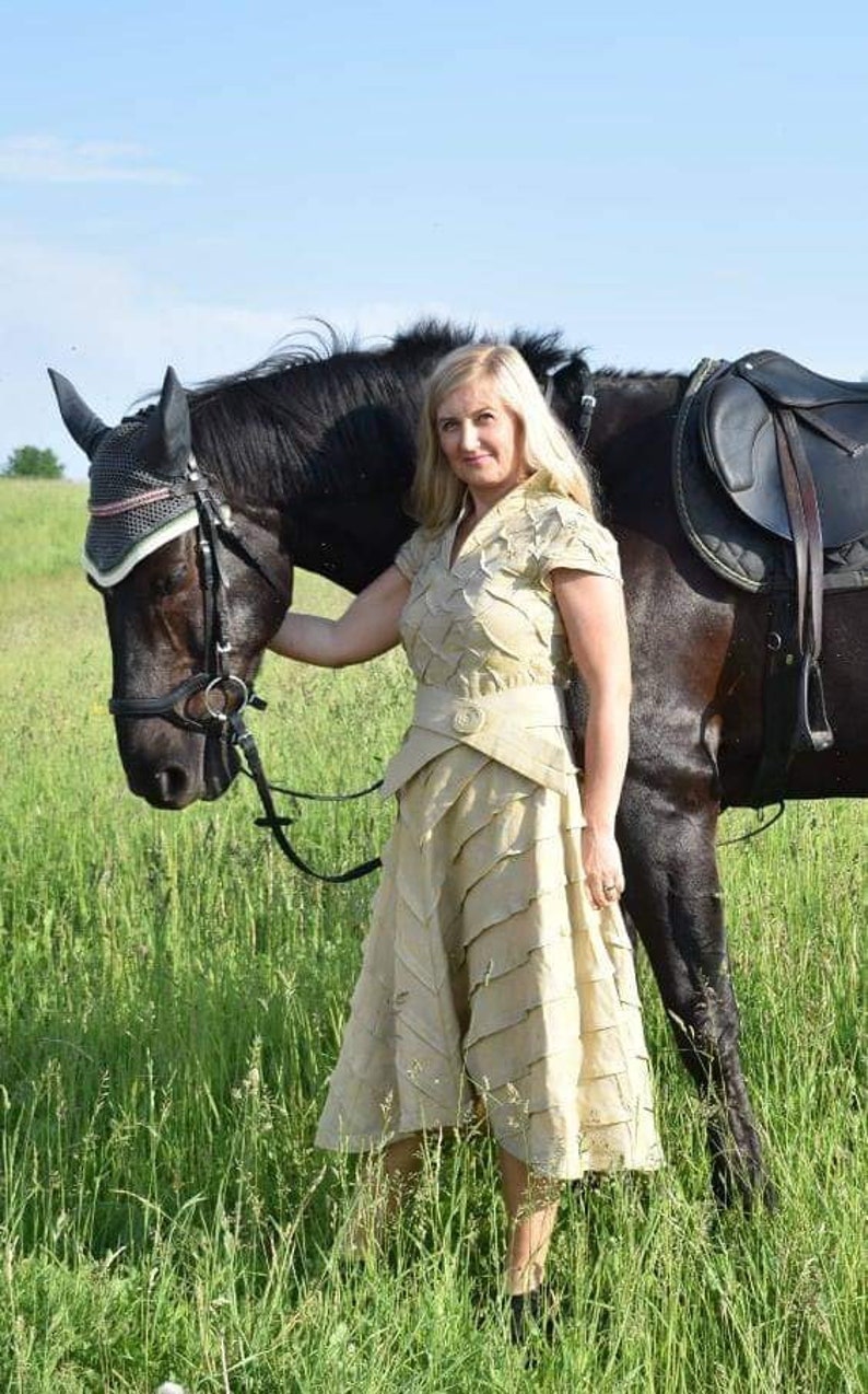 May include: A woman in a beige dress stands next to a black horse in a grassy field. The dress has a layered design and a matching belt. The horse wears a bridle and saddle, suggesting equestrian activities.