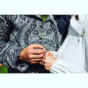 Puede incluir: Primer plano de una pareja intercambiando anillos durante una ceremonia de boda. El novio lleva una camisa de paisley blanco y negro, mientras que la novia lleva un vestido blanco.