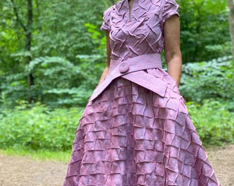 May include: A woman wearing a long, pink dress with a textured, quilted pattern. The dress has a high collar and short sleeves. The woman is standing in a forest setting.
