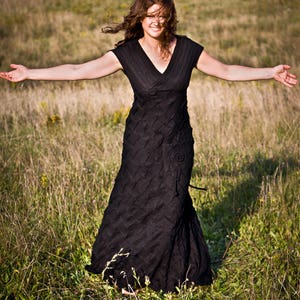 May include: A woman wearing a black dress stands in a field of long grass with her arms outstretched. She is smiling and her hair is blowing in the wind.