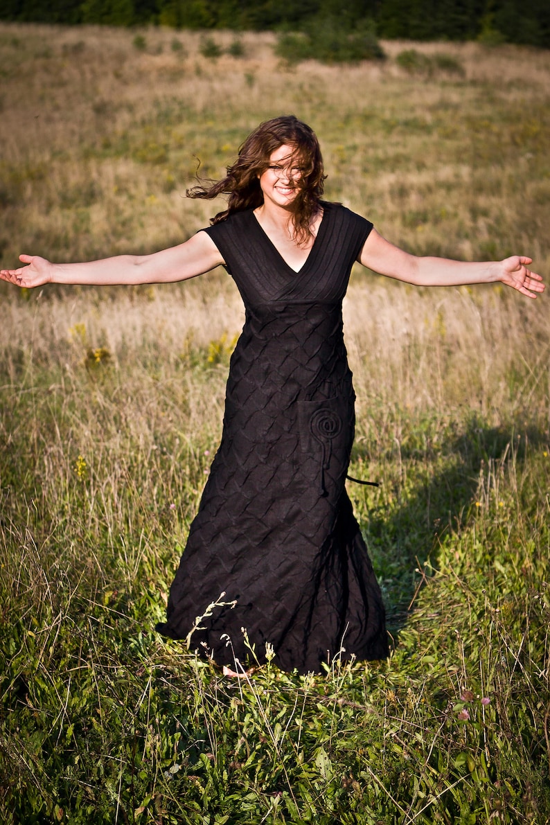 May include: A woman wearing a black dress stands in a field of long grass with her arms outstretched. She is smiling and her hair is blowing in the wind.