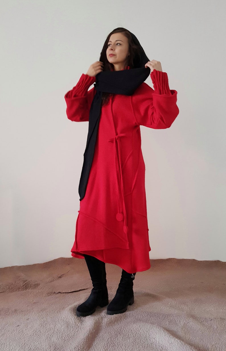 May include: A red hooded dress with a black scarf. The dress has a long, flowing skirt and a cinched waist. The model is wearing black boots.