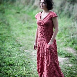 May include: A woman wearing a long, rust-coloured dress with a textured pattern stands in a grassy field. The dress has a V-neckline and short sleeves. She is looking off to the side.