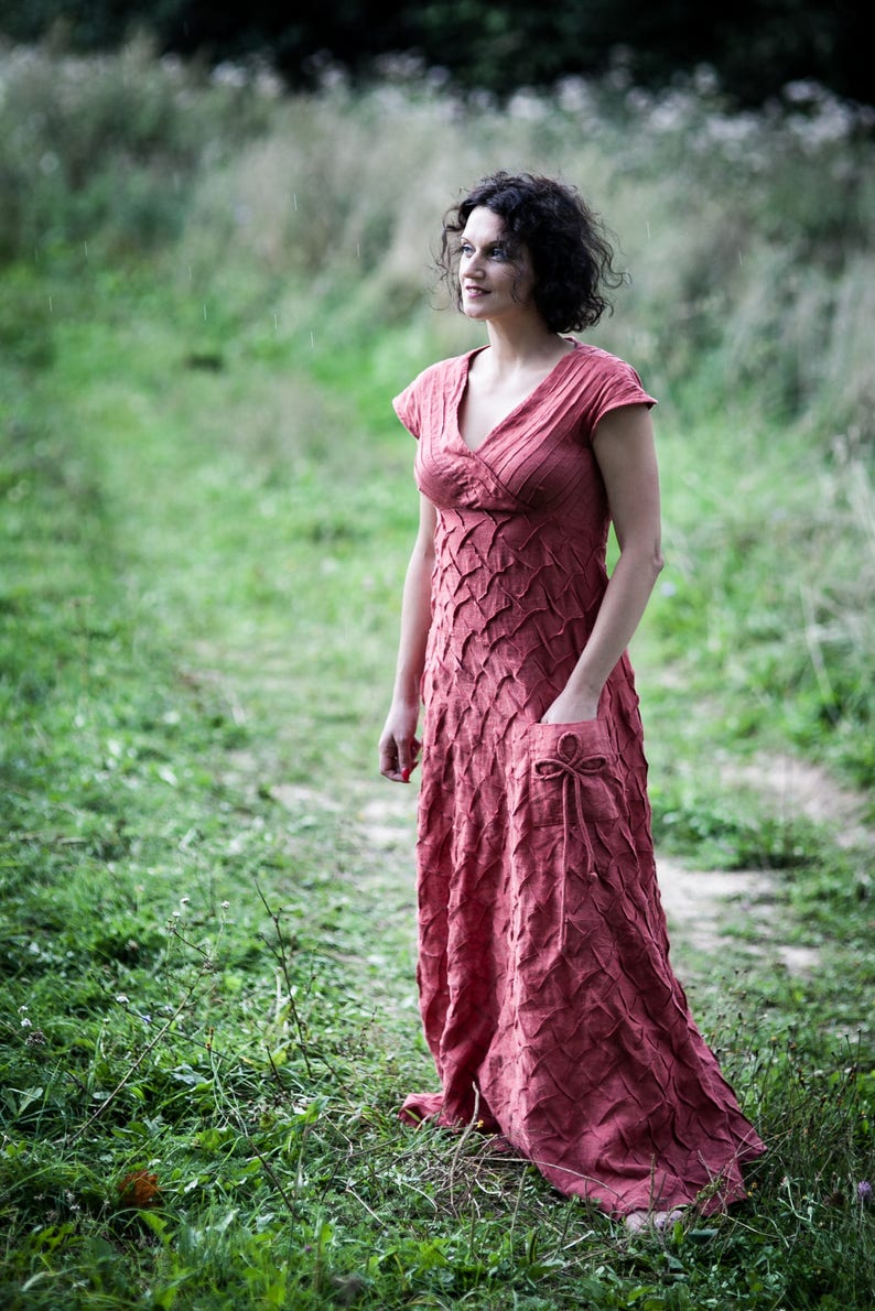May include: A woman wearing a long, rust-coloured dress with a textured pattern stands in a grassy field. The dress has a V-neckline and short sleeves. She is looking off to the side.