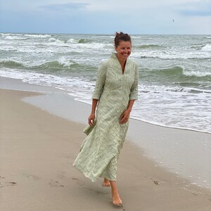 May include: A woman in a light green dress walks on a sandy beach with waves crashing in the background.