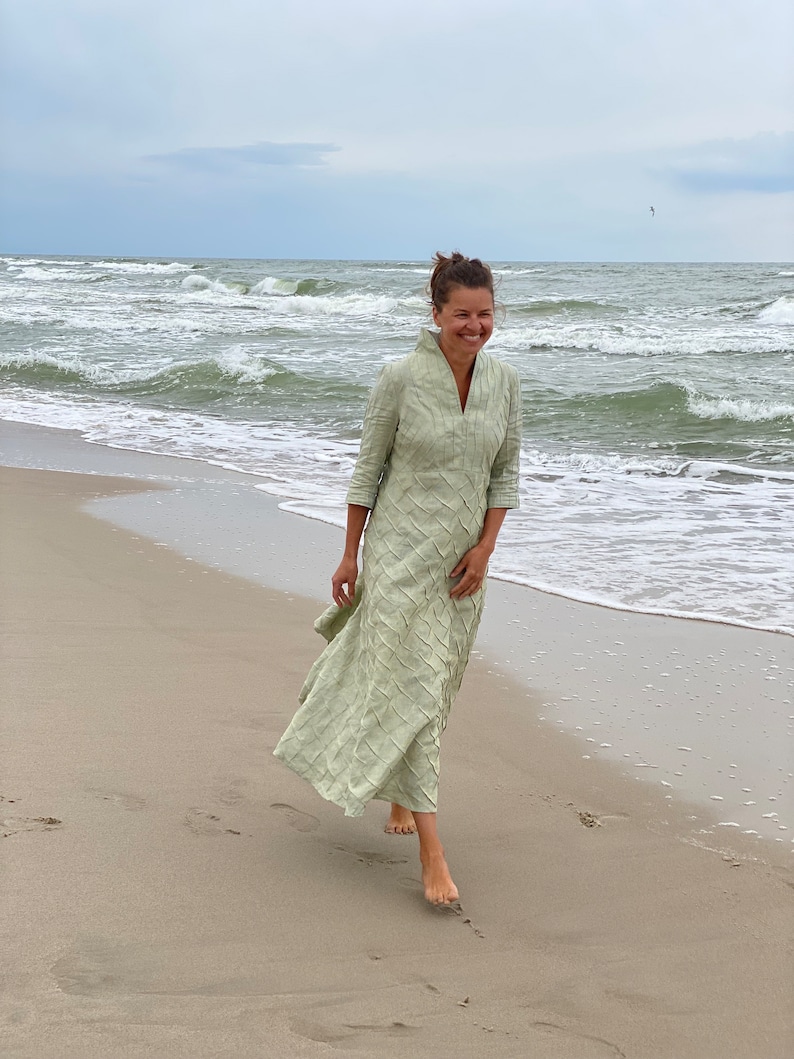 May include: A woman in a light green dress walks on a sandy beach with waves crashing in the background.