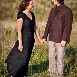 May include: A couple standing in a field, holding hands and looking at each other. The woman is wearing a black dress and the man is wearing a brown shirt and beige trousers.