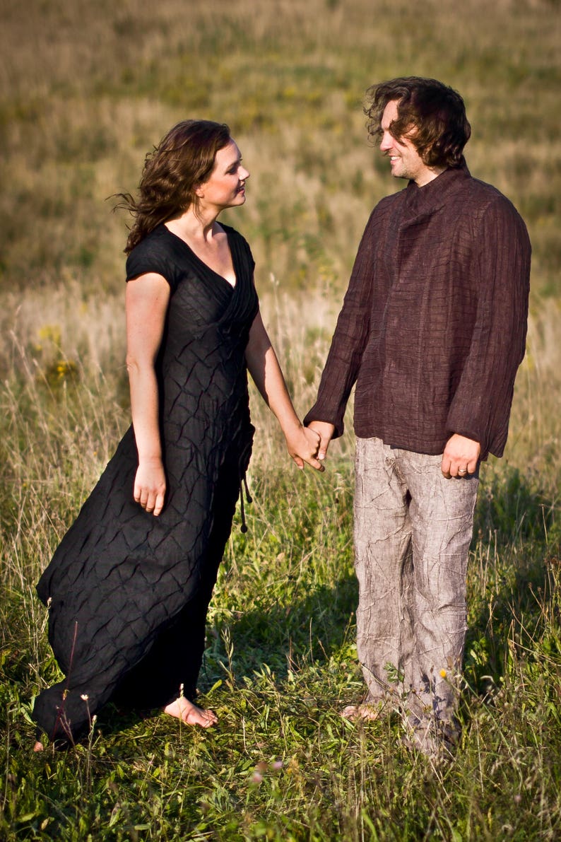 May include: A couple standing in a field, holding hands and looking at each other. The woman is wearing a black dress and the man is wearing a brown shirt and beige trousers.