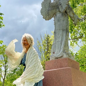 Puede incluir: Una mujer con un chal blanco se encuentra de pie frente a una estatua de &aacute;ngel de piedra. La mujer sonr&iacute;e y el chal ondea con el viento. La estatua est&aacute; sobre un pedestal de piedra.