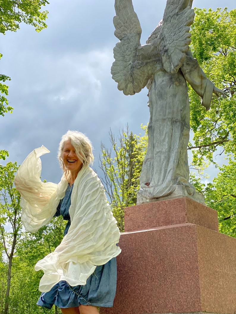Puede incluir: Una mujer con un chal blanco se encuentra de pie frente a una estatua de &aacute;ngel de piedra. La mujer sonr&iacute;e y el chal ondea con el viento. La estatua est&aacute; sobre un pedestal de piedra.