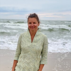 May include: A woman wearing a light green, textured linen dress stands on a sandy beach with the ocean in the background.