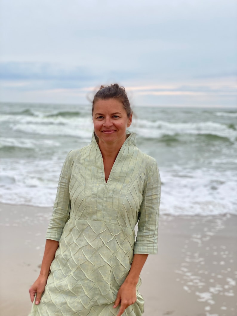 May include: A woman wearing a light green, textured linen dress stands on a sandy beach with the ocean in the background.