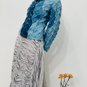 May include: A blue and teal textured jacket with a button-up front and a high collar. The jacket is worn over a grey skirt.