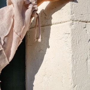 May include: A close-up of a light pink linen blouse with a gathered sleeve detail and a drawstring tie. The sleeve is gathered at the wrist and has a small, round, decorative button.