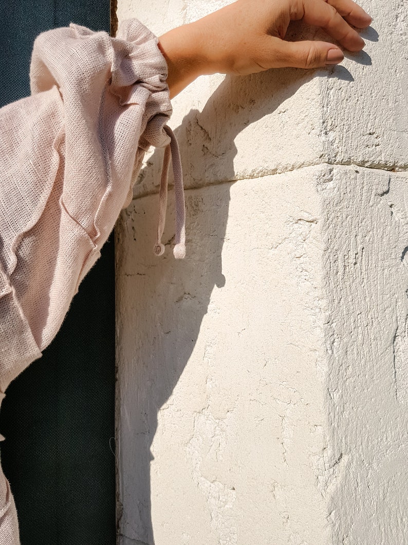 May include: A close-up of a light pink linen blouse with a gathered sleeve detail and a drawstring tie. The sleeve is gathered at the wrist and has a small, round, decorative button.