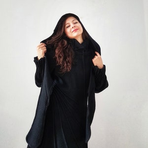 May include: A woman wearing a black hooded coat with a long, flowing design. The coat has a high collar and a relaxed fit.