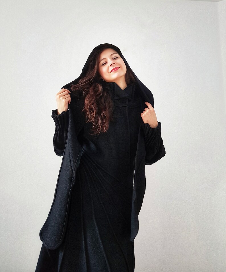 May include: A woman wearing a black hooded coat with a long, flowing design. The coat has a high collar and a relaxed fit.