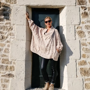May include: A woman wearing a light pink, textured, oversized sweater with a cowl neck and black leather pants. She is standing in a doorway with a stone wall behind her. She is wearing sunglasses and brown sneakers.
